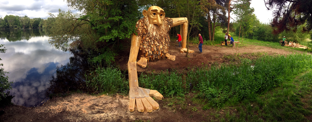 Teddy Friendly is made from local scrapwood, including cut offs from cut down local trees, which has been used for his fur. He is standing at a water stream, at a lake holding out his hand helping people to cross (click on the “Source” of any image to see a gallery of that sculpture).